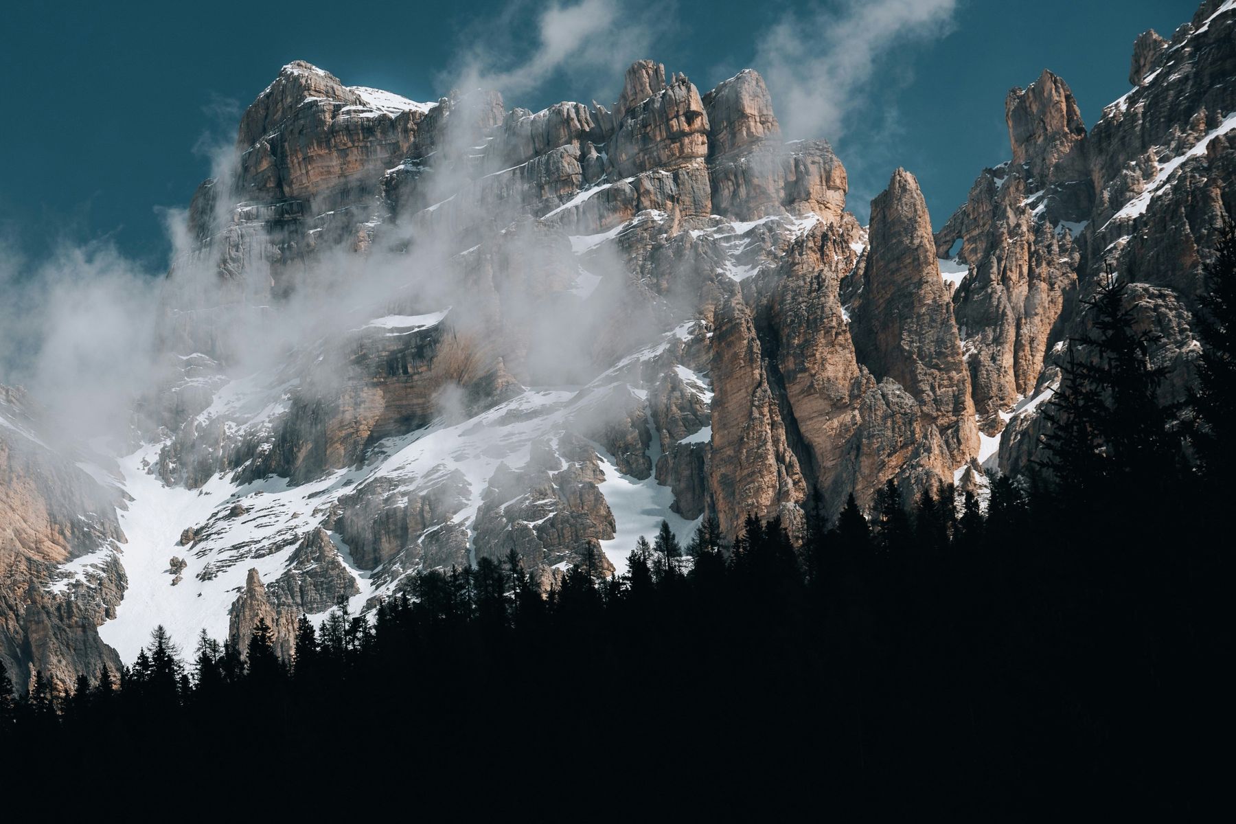 Tall rock faces with snow and drifting mist above a silhouette of forest