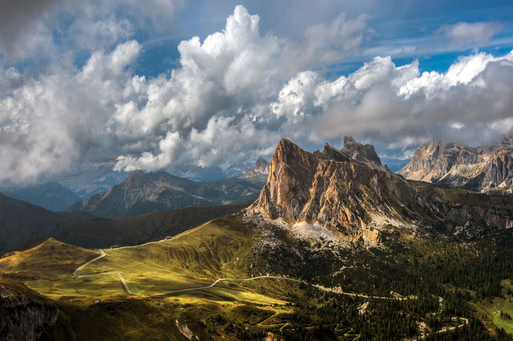 A winding pass road below a rocky peak and big clouds