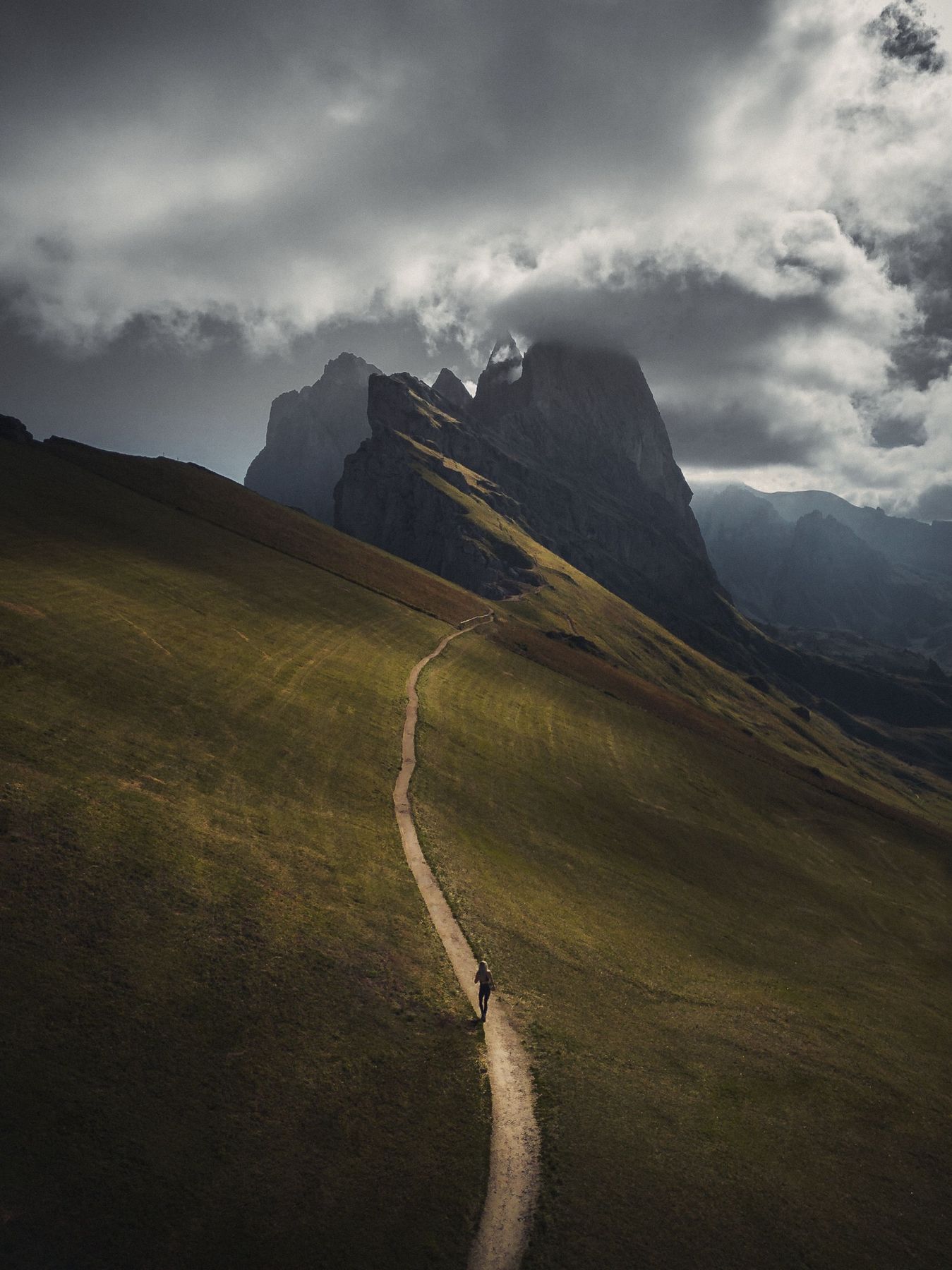 A lone hiker on a winding path across a green slope below a cloud-covered Seceda ridge