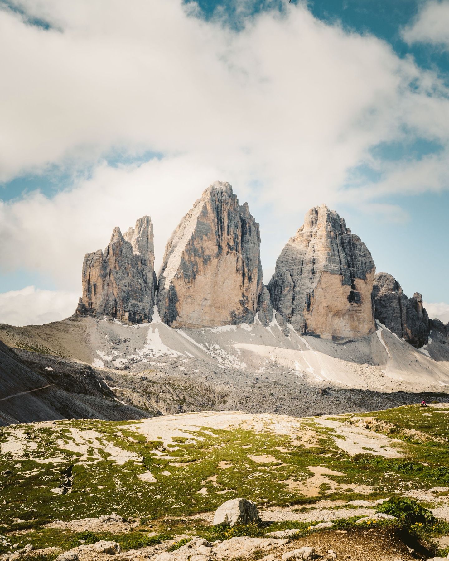 The three rock towers of Tre Cime di Lavaredo rising above green and white rocky ground under big clouds