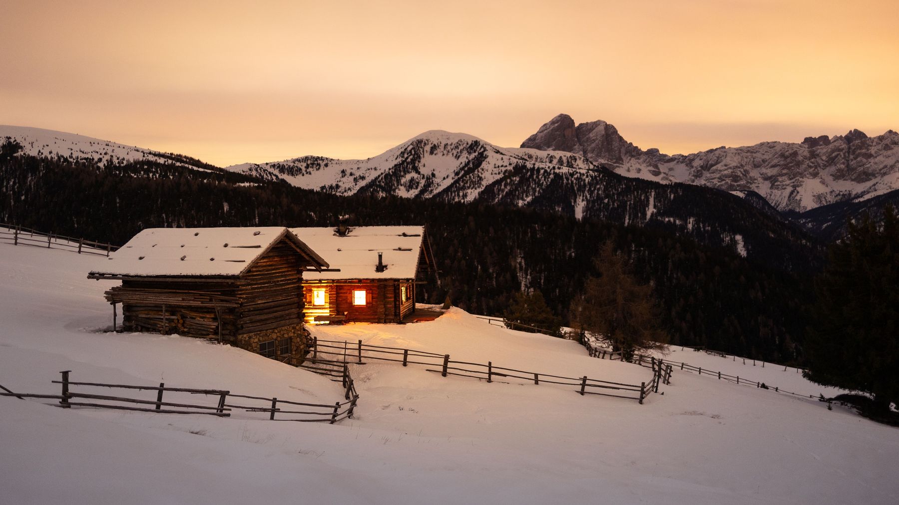 A lit log cabin in deep snow at sunset with snowy peaks behind