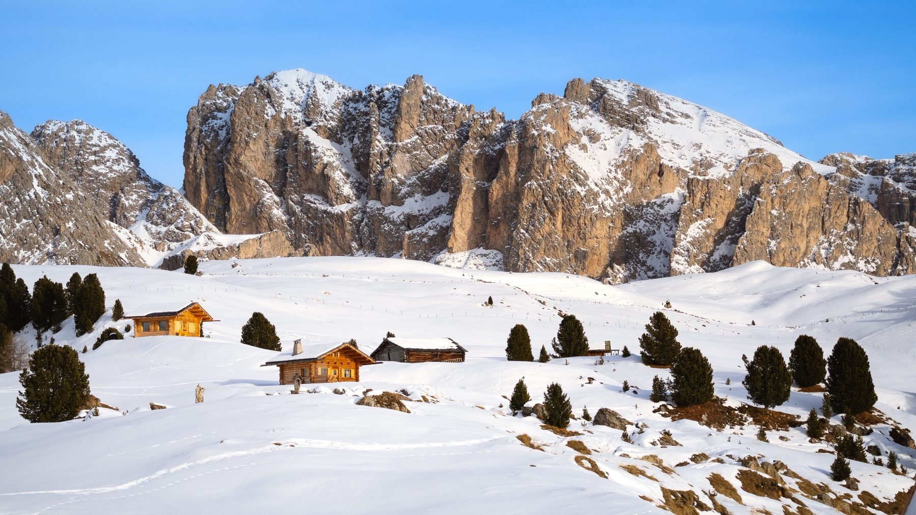 Wooden huts in a sunny snowy meadow below pale rock walls